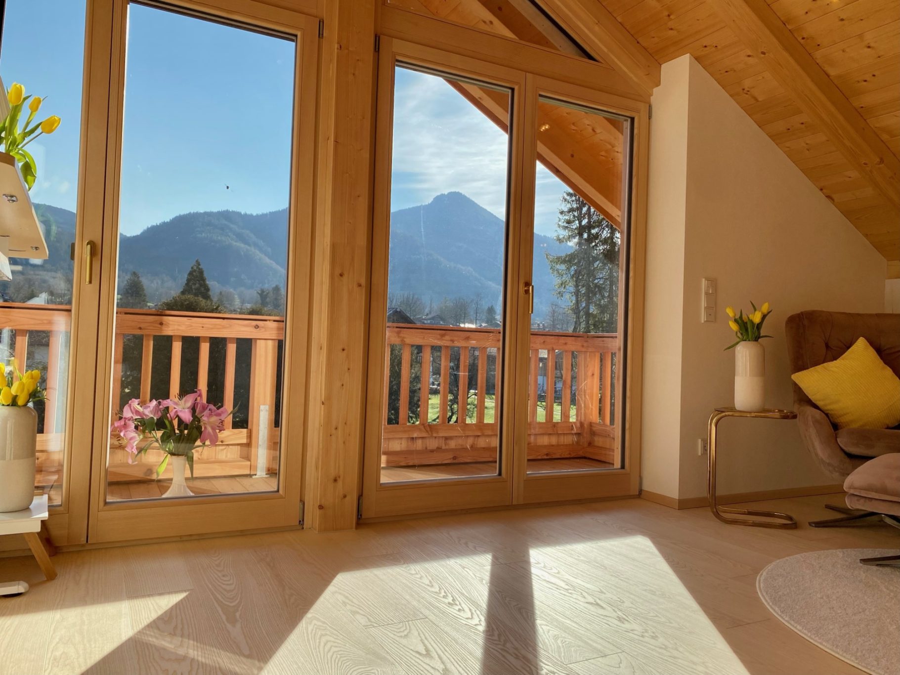 Heller Wohnraum mit großen Fenstern, Blick auf Berge und bunten Blumen im Vordergrund.
