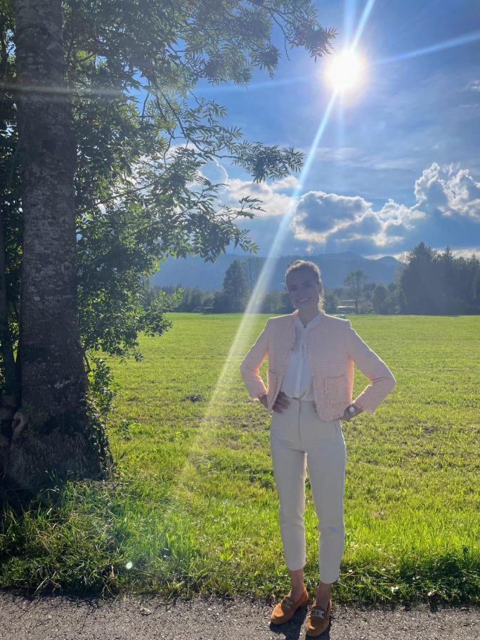 Person steht im Freien auf einer Wiese, umgeben von Bäumen und unter blauem Himmel.